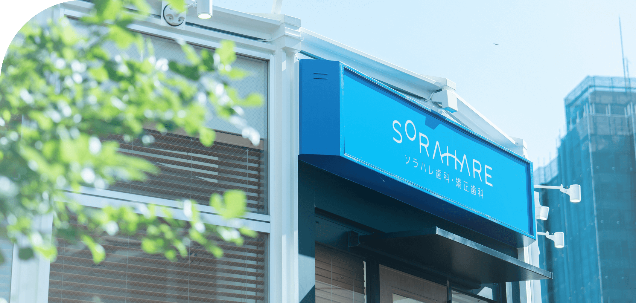 東大和市・玉川上水駅から徒歩1分にあるソラハレ歯科・矯正歯科の外観。晴れた空の下、緑の木々の隣に立つモダンでクリーンな建物。